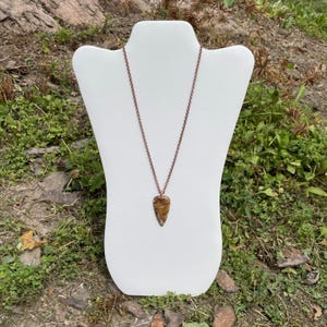 Op de afbeelding: Een koperen ketting met een bruine en gouden pijlpunt hanger. De ketting wordt getoond op een witte mannequinbuste.