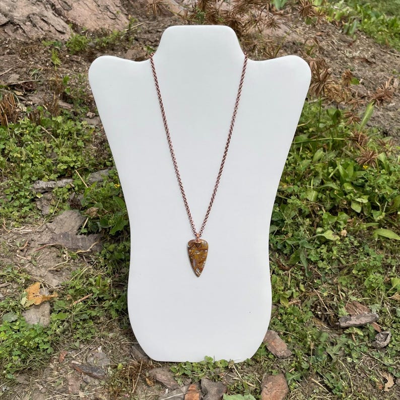 Op de afbeelding: Een koperen ketting met een bruine en gouden pijlpunt hanger. De ketting wordt getoond op een witte mannequinbuste.