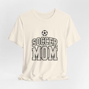 May include: A cream-colored short-sleeve t-shirt featuring a black and white graphic on the chest. The graphic includes a soccer ball icon positioned above the text SOCCER MOM, which is written in a bold, block-style font with a dotted pattern.