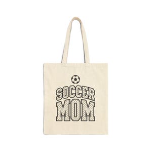 May include: A beige canvas tote bag featuring a black graphic print of a soccer ball centered above the text SOCCER MOM in a bold, dotted varsity-style font. The bag has two long shoulder straps and a rectangular body made of fabric.
