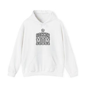May include: A white hooded sweatshirt featuring a front kangaroo pocket and long sleeves. The center of the chest displays a black and white graphic of a soccer ball above the text SOCCER MOM in a bold, block-style font with a dotted pattern.