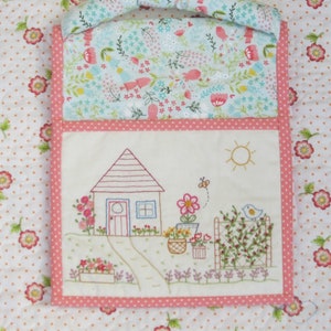 Könnte beinhalten: Ein rosa-weißer gesteppter Wandbehang mit einem handgestickten Design eines Hauses mit Garten. Das Haus hat ein rosafarbenes Dach, eine weiße Tür und zwei Fenster. Der Garten hat einen Zaun, Blumen, einen Vogel und eine Sonne.