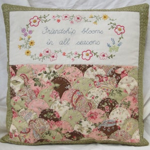 May include: A white pillow with a green border and a floral embroidered design. The embroidery includes the text "Friendship blooms in all seasons". The back of the pillow is made of a patchwork of fabric in shades of pink, green, and brown.
