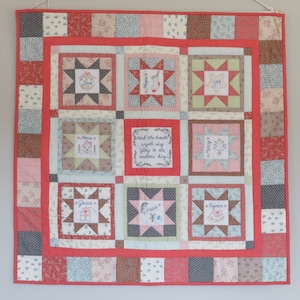 May include: A red, white, and blue patchwork quilt with a Christmas theme. Each square features a different design, including stars, ornaments, and a house. The text "Hear the herald angels sing 'Glory to the newborn King!" is embroidered in one of the squares.