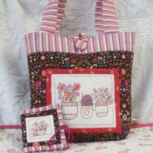 May include: A floral tote bag and matching small notebook. The tote bag features a black background with a colorful floral pattern, a red border, and a white square with embroidered flowers in pots. The handles and notebook are striped.
