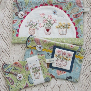 Puede incluir: Tres pequeñas bolsas con cremallera con un patrón de paisley y flores bordadas en macetas. Las bolsas tienen un esquema de color azul, verde y blanco. La bolsa más grande tiene un fondo blanco con un diseño de medio círculo y el texto "HAPPY".