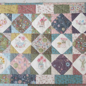 May include: A patchwork quilt featuring a range of floral and nature-inspired embroidery designs. The quilt is composed of squares in various colours and patterns, with the embroidery worked in a variety of colours. The quilt is bordered with a brown fabric.