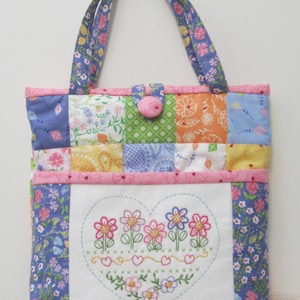 May include: A patchwork tote bag with a floral design. The bag has a heart-shaped embroidered patch with flowers and a row of hearts. The bag has a pink button closure.