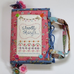 May include: A pink and blue fabric book with a handle. The book has a white patch with the words "Wooly Things..." embroidered on it. The book is decorated with buttons and tassels.