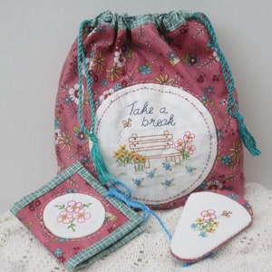 Könnte beinhalten: Eine rosa Stofftasche mit Kordelzug und türkisfarbenem Besatz und einem weißen gestickten Kreis, auf dem "Take a break" mit einer Bank und Blumen steht. Es gibt zwei kleinere passende Taschen mit gestickten Blumen.