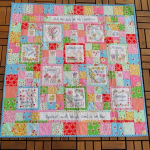 Może przedstawiać: Kolorowa patchworkowa kołdra z niebieską obwódką. Kołdra zawiera kwadraty z różnych tkanin w kwiatowe wzory. Haftowane kwadraty wyświetlają słowa takie jak "pokój", "radość" i "dobroć", wraz z kwiatowymi wzorami i tekstem z Listu do Galatów.