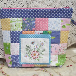 Puede incluir: Una bolsa con cremallera de patchwork con un dise&ntilde;o floral azul, rosa y verde. La bolsa tiene un peque&ntilde;o bolsillo bordado con un dise&ntilde;o floral y una mariposa.