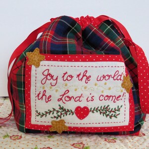Könnte beinhalten: Rote und grüne karierte Kordeltasche mit einem weißen bestickten Feld, auf dem "Joy to the world the Lord is come!" mit goldenen Sternen und einem roten Herzen steht.