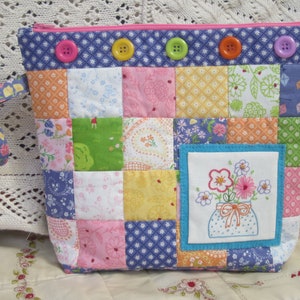 Puede incluir: Una bolsa de cremallera de tela patchwork con un diseño bordado floral azul y blanco. La bolsa tiene un diseño patchwork rosa, azul y amarillo con una cremallera azul y un asa de tela azul.