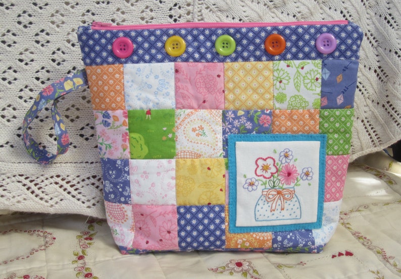 Puede incluir: Una bolsa de cremallera de tela patchwork con un dise&ntilde;o bordado floral azul y blanco. La bolsa tiene un dise&ntilde;o patchwork rosa, azul y amarillo con una cremallera azul y un asa de tela azul.