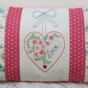 Peut inclure: Un oreiller blanc avec des côtés en tissu à pois roses et blancs. Le devant de l'oreiller est blanc avec un cœur brodé rose et vert avec le mot "love" à l'intérieur. L'oreiller est fait de tissu patchwork avec une variété de motifs floraux.