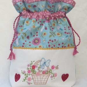Może przedstawiać: Biała torba ze sznurkiem do ściągania z niebieskim i białym nadrukiem kwiatowym. Torba ma białą podszewkę z haftowanym wzorem koszyka z kwiatami i niebieską kokardką.