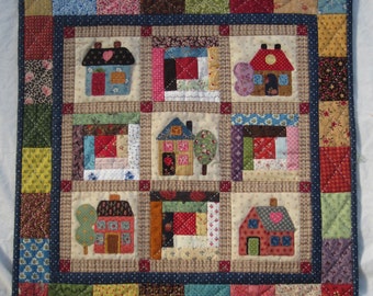 May include: A colorful patchwork quilt with a central design of nine squares featuring houses with different colors and patterns. The quilt is framed with a dark blue border and a colorful outer border.