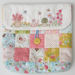 May include: A white fabric pouch with a patchwork design. The pouch has a flap with embroidered flowers, a cat, and butterflies. The flap is closed with a yellow button and blue string.