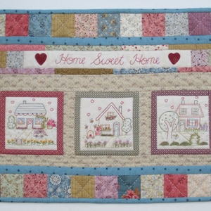 Puede incluir: Un tapiz de pared acolchado con fondo beige y borde azul. El centro presenta tres imágenes bordadas de casas con techos rojos y árboles verdes. La parte superior del edredón tiene una pancarta con el texto "Home Sweet Home" en bordado rojo.