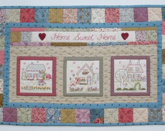 May include: A quilted wall hanging with a beige background and a blue border. The center features three embroidered images of houses with red roofs and green trees. The top of the quilt has a banner with the text "Home Sweet Home" in red embroidery.