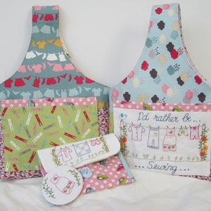 May include: Two blue and white fabric tote bags with a green and white fabric pocket on the front. The tote bags have a handle and are decorated with a sewing theme. One bag has the text "I'd rather be... Sewing..." embroidered on the front. The other bag has a pocket with a green and white fabric with a pattern of clothespins and clotheslines.