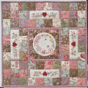 Könnte beinhalten: Eine quadratische Patchwork-Decke mit einem zentralen, runden Stickmuster, das Blumen und eine Biene zeigt. Die Decke besteht aus verschiedenen floralen Stoffen in den Farben Rosa, Braun und Grün.