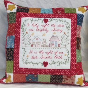 Könnte beinhalten: Ein rotes und weißes Weihnachtskissen mit einem gesteppten Patchwork-Muster. Das Kissen zeigt eine gestickte Szene mit Häusern, Bäumen und Sternen mit dem Text "O holy night the stars are brightly shining. It is the night of our dear Savior's birth."