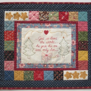 May include: A quilted wall hanging with a patchwork design in red, green, blue, and yellow. The centre features a white panel with embroidered text: "...God so loved the world... he gave his one and only Son..." with two stylised Christmas trees and a gift box.