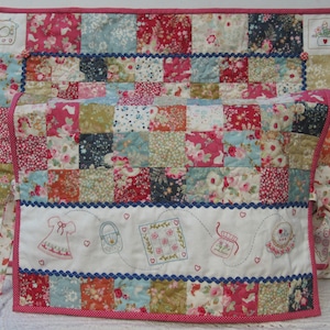 May include: A patchwork quilt with a floral design in shades of pink, red, blue, and green. The quilt has a white border with embroidered images of a sewing machine, a dress, a purse, a watering can, and a sewing basket. The quilt is tied with pink ribbon.