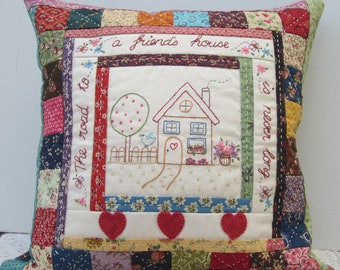 May include: A white pillow with a patchwork design featuring a house with a red heart on the door and the text "a friend's house is never too far to go".