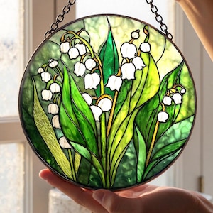 Puede incluir: Un panel de vidriera redondo con un diseño de lirios del valle. La obra de arte muestra flores blancas en forma de campana, hojas verdes y una cadena para colgar. El vidrio tiene una variedad de tonos verdes y amarillos.