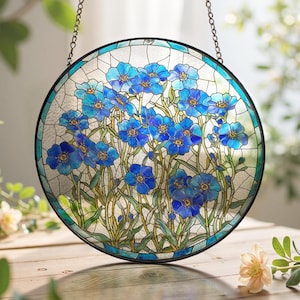 May include: Circular stained glass window with a black frame and hanging chain. The design features blue flowers with yellow centres and green stems. The border is teal. The window is approximately 25 cm in diameter.