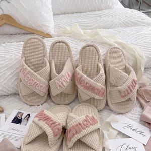 May include: Six beige waffle-textured slippers with crisscross straps featuring embroidered names in pink thread. The names include Bridesmaid, Aria, Maid of Honor, Luna, Layla, and Amelia. Each slipper has a flat sole and open-toe design.