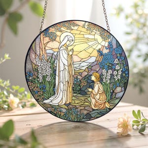 Peut inclure: Vitrail rond repr&eacute;sentant une sc&egrave;ne religieuse. Une femme en robe blanche se tient devant une personne agenouill&eacute;e, entour&eacute;e de fleurs et d'arbres. Le soleil brille vivement en arri&egrave;re-plan, avec un cadre en m&eacute;tal noir et une cha&icirc;ne de suspension.