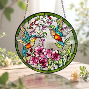 Op de afbeelding: Een rond gebrandschilderd glazen raam met twee kolibries met kleurrijke vleugels, te midden van roze bloemen en groene bladeren. Het raam heeft een groene rand en hangt aan een ketting.