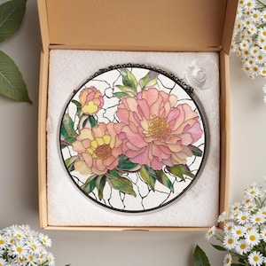 May include: Round stained glass window art featuring pink and yellow peony flowers with green leaves. The floral design is set against a white background with black outlines. The artwork is displayed in a cardboard box with a suction cup attached.