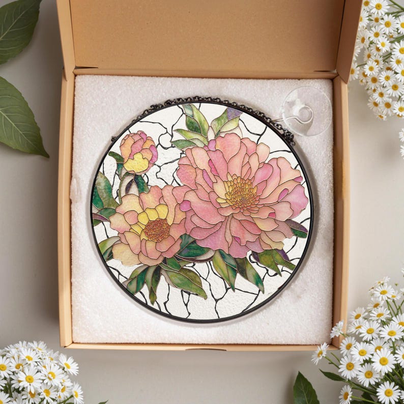 May include: Round stained glass window art featuring pink and yellow peony flowers with green leaves. The floral design is set against a white background with black outlines. The artwork is displayed in a cardboard box with a suction cup attached.
