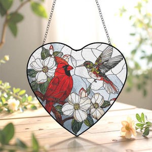 May include: Heart-shaped stained glass window art featuring a vibrant red cardinal and a hummingbird. The design includes white flowers and a blue background, all framed with a black border and suspended by a chain.