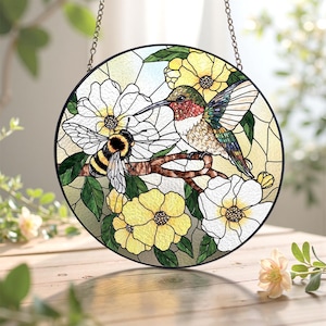 May include: Round stained glass window featuring a hummingbird and a bee perched on a branch with white and yellow flowers. The hummingbird has iridescent feathers, and the bee has black and yellow stripes. The window is framed in black and hangs from a chain.