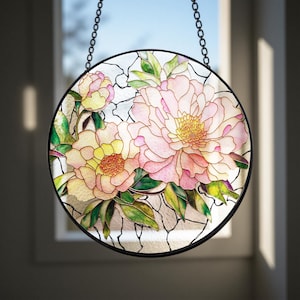 May include: Round stained glass window art featuring a floral design with pink and yellow peonies and green leaves. The glass is framed in black and hangs from a chain. The window art is a decorative home decor item.