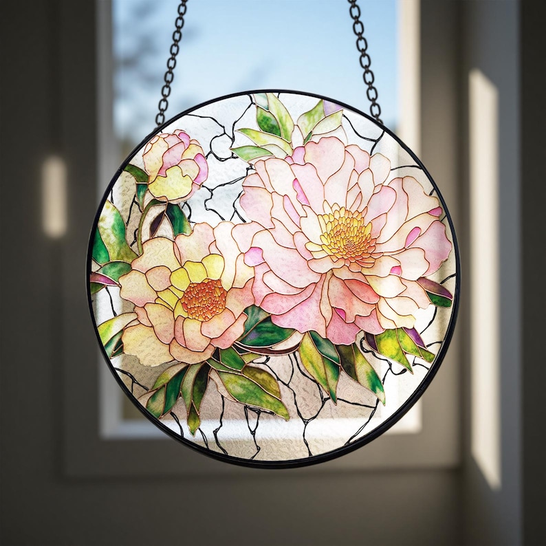 May include: Round stained glass window art featuring a floral design with pink and yellow peonies and green leaves. The glass is framed in black and hangs from a chain. The window art is a decorative home decor item.