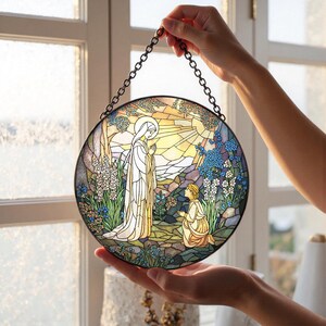 Peut inclure: Panneau de vitrail rond repr&eacute;sentant une sc&egrave;ne religieuse avec une femme en robe blanche et une figure agenouill&eacute;e. L'&oelig;uvre d'art pr&eacute;sente une palette de couleurs vives avec des bleus, des verts et des jaunes, et est encadr&eacute;e d'une cha&icirc;ne en m&eacute;tal noir.