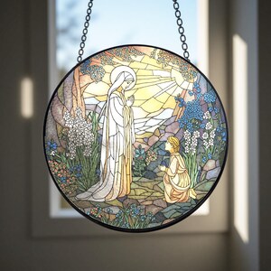 Peut inclure: Panneau de vitrail rond repr&eacute;sentant une sc&egrave;ne religieuse. Une femme en robe blanche se tient debout, les mains jointes, face &agrave; une personne agenouill&eacute;e. L'arri&egrave;re-plan pr&eacute;sente un soleil, des arbres et des fleurs dans des tons de bleu, jaune et vert. Le panneau est encadr&eacute; en noir et suspendu &agrave; une cha&icirc;ne.
