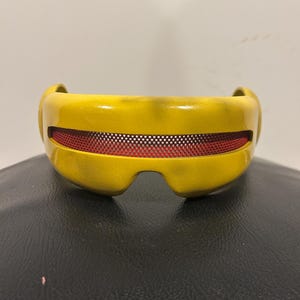 May include: A yellow visor with a smooth, curved frame and a horizontal slit across the front. The slit features a fine red mesh screen. The yellow surface has subtle, darker shading, and the design includes a molded nose bridge cutout.