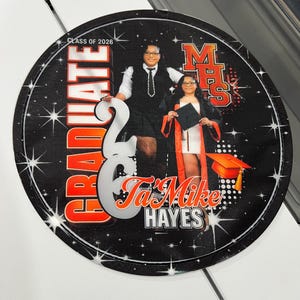 May include: A round, black graduation keepsake featuring two people in graduation attire. The text "GRADUATE" and "CLASS OF 2026" are in orange and white. The name "Ta'Mike HAYES" is also displayed.
