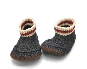 Puede incluir: Un par de zapatillas de crochet grises con un puño a rayas blancas y rojas. Las zapatillas tienen una suela de cuero marrón.