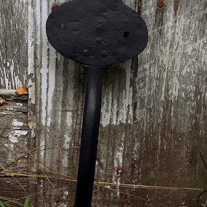 May include: A black, decorative metal door handle with a unique shape. The handle has a long, straight stem connecting two rounded, flat shapes, with a triangular top. The handle is mounted on a weathered wooden door.