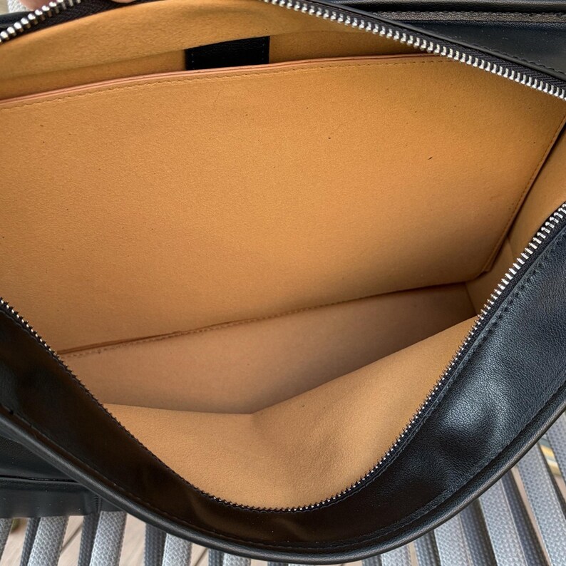 May include: Open view of a black leather briefcase with a silver zipper. The interior is tan with a small pocket. The briefcase is open, revealing the inside lining and construction.