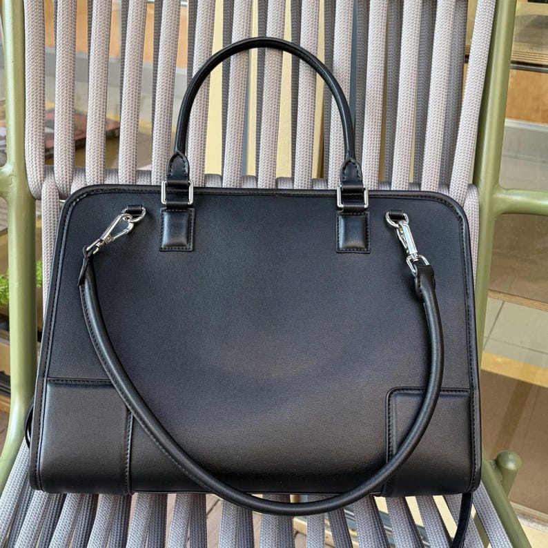 May include: Black leather briefcase with a top handle and a detachable shoulder strap. The bag has silver-tone hardware and a rectangular shape. The briefcase is sitting on a gray and green chair.